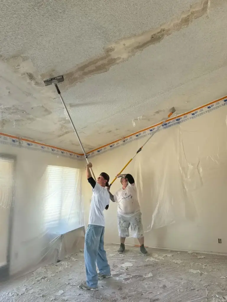 Two workers removing popcorn ceiling texture with pole scrapers in a plastic-protected room