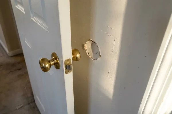 Open bedroom door with knob aligned to a round drywall crater at handle height, lit with raking window light